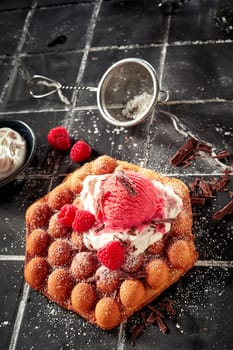 Tasty fresh raspberry and bubble waffle summer dessert with berry ice-cream on a bed of whipped cream sprinkled with chocolate flakes in a high angle vertical view on tiles with copyspace