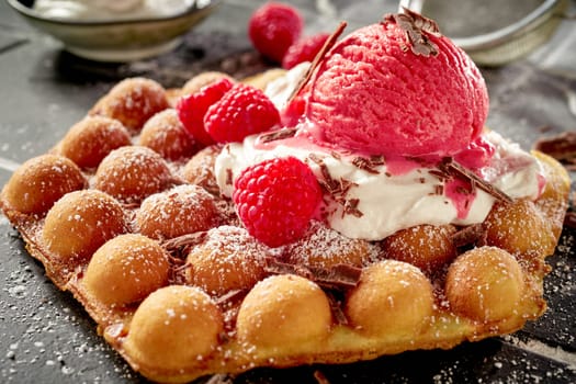 Traditional egg waffle or bubble waffle topped with tasty raspberry ice cream and garnished with fresh fruit on whipped cream for a delicious gourmet summer dessert