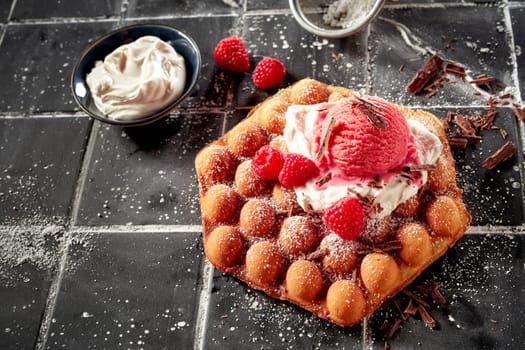 Hexagonal crispy golden bubble waffle or egg waffle topped with whipped cream, berry ice-cream and fresh tropical raspberries sprinkled with flaked chocolate in high angle with copyspace for menu advertising