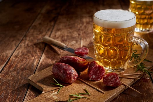 Tasty spicy mini salami served with a cold tankard of beer and fresh rosemary on a rustic cutting board on a wooden table with copyspace for advertising