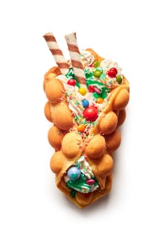 Top view of egg waffle filled with ice cream and multicolored candies and sprinkles with waffle rolls with chocolate on white background