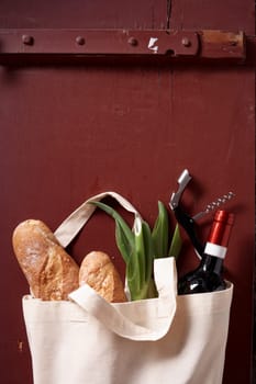Top view of eco friendly textile bag with groceries and botte of wine placed near corkscrew on red wooden table