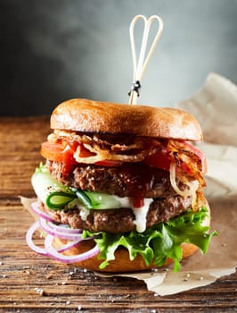 Appetizing double burger with vegetables and caramelized onions served on parchment paper on wooden table against gray background in light studio