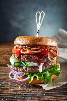 Appetizing burger with cutlets and vegetables and fried onion served on parchment paper on wooden table in room on gray background