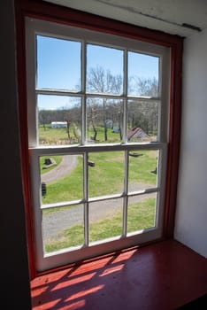 Colonial American rural village lane and buildings seen through an antique red 12-pane Pennsylvania window with sunlight streaming in. Copy space, no people.
