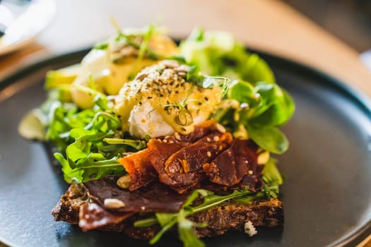 Eggs benedict with dried ham and salad mix on a black plate. A view of the closeness.