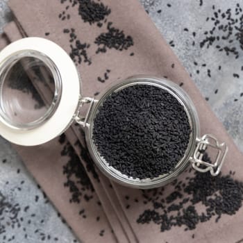 Indian spice Black cumin (nigella sativa or kalonji) seeds glass jar on grey table top view. Traditional asian medicine, healthy and vegetarian food concept