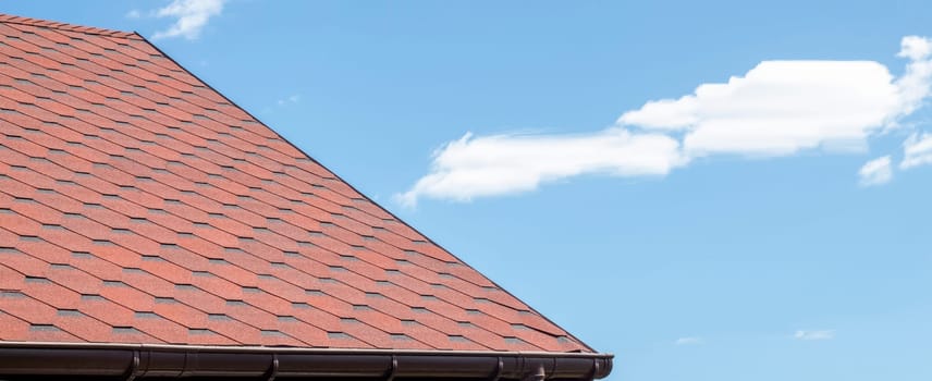 New roof with red shingles against the blue sky. Tiles on the roof of the house. Use to advertise roof fabrication and maintenance. Spotted texture. Affordable roofing. Banner with copy space