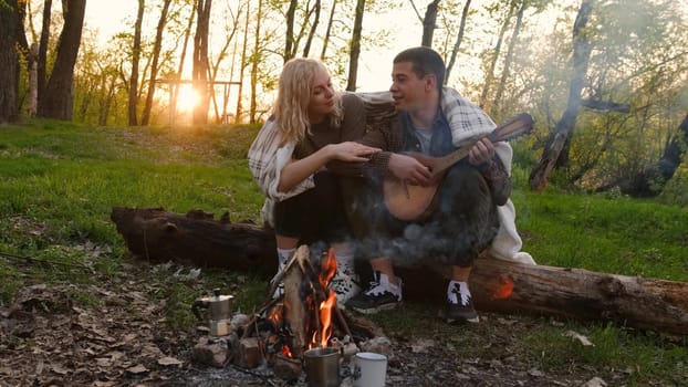 Closeup portrait of a couple sitting with guitar near bonfire in the forest.