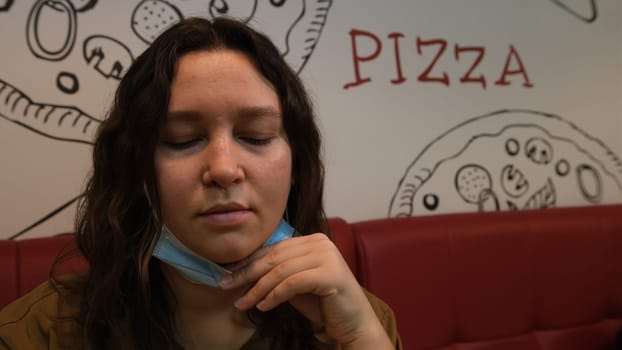 Beautiful young woman in mask eating pizza in the restaurant. Happy and joyful lifestyle. Healthy and junk food concept. Vegetarian food. Vegetarian pizza. Covid 19, Coronavirus.