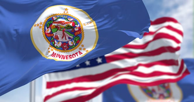The Minnesota state flag waving along with the national flag of the United States of America. In the background there is a clear sky. Minnesota is a state in the upper Midwestern United States