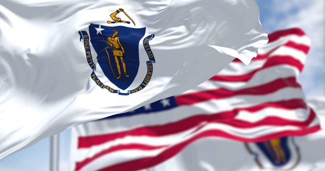 The Massachusetts state flag waving along with the national flag of the United States of America. In the background there is a clear sky. Massachusetts is a state in the New England region of the United States