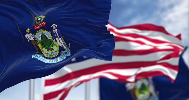 The Maine state flag waving along with the national flag of the United States of America. In the background there is a clear sky. Maine is a state in the New England region of the United States