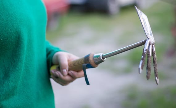 Caucasian woman gardener in the garden shows a rake. The concept of gardening and sustainable summer. Woman's hand with a garden tool. Chopper rake for gardening in female hands. Place for text