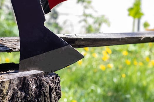 A new modern ax with a red handle sticks out of a wooden stump against a green meadow on a summer sunny day. The ax stuck into the stump. Ax blade in a log