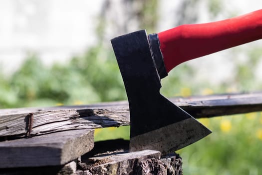 A new modern ax with a red handle sticks out of a wooden stump against a green meadow on a summer sunny day. The ax stuck into the stump. Ax blade in a log