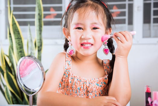 Asian adorable funny little girl making makeup her face she look in the mirror and cheek fluffy brush for powder, Learning activity to be woman, happy kid is beautiful make up with cosmetics toy