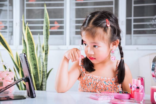 Asian funny little girl making makeup her face she look in smartphone and use eyebrow mascara, Learning activity to be woman, social media blogger recording vlog with cosmetics, online tutorial