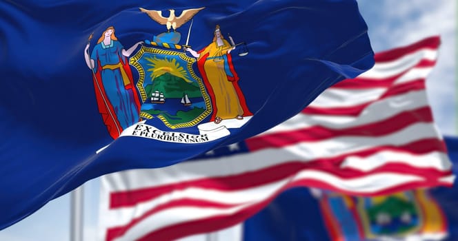 The New York state flag waving along with the national flag of the United States of America. In the background there is a clear sky. New York is a state in the Northeastern United States