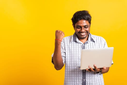 Closeup Asian happy portrait young black man excited holding laptop computer clenching fists and raising a hand for winner sign celebrating his success, studio shot isolated on yellow background