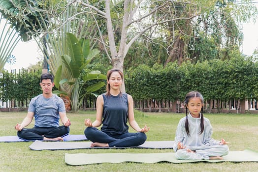 Asian young mother, father practicing doing yoga exercises with child daughter outdoors in meditate pose together in nature a field garden park, family kid sport and exercises for healthy lifestyle