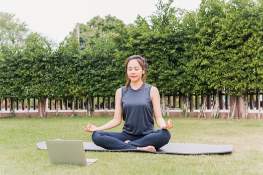 Asian young woman using laptop during online video training practicing yoga outdoors in meditate pose sitting on green grass in nature a field garden park, Stretching health care concept