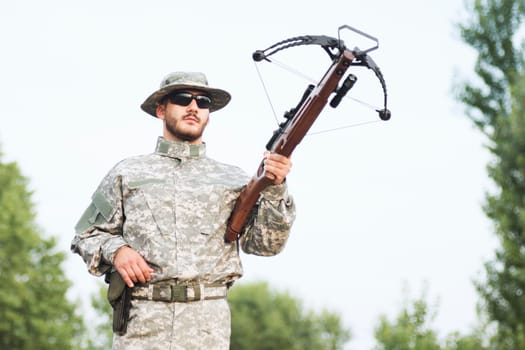 The hunter or army soldier in military uniform is holding a crossbow weapon outdoors.