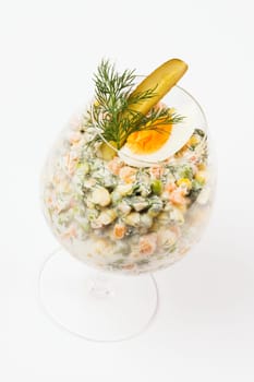 Russian salad with garnish is served in drinking glass on white background.