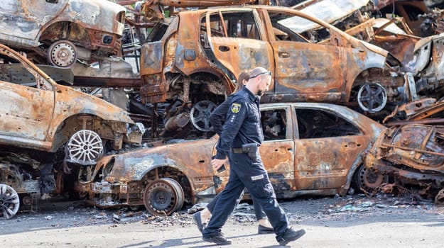 Police officers in front of burnt cars destroyed by rocket explosions. War in Ukraine. Destroyed vehicles of civilians. Car graveyard. Ukraine, Irpen - May 12, 2022