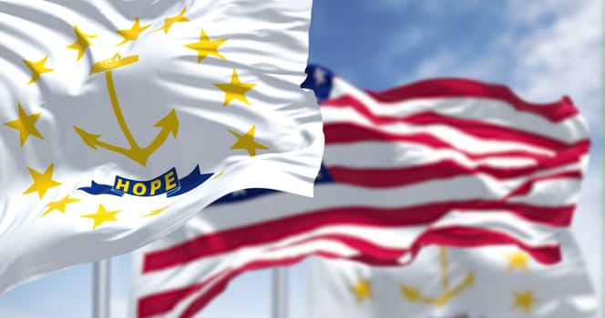 The Rhode Island state flag waving along with the national flag of the United States of America. In the background there is a clear sky. Rhode island is a state in the the Northeastern United States