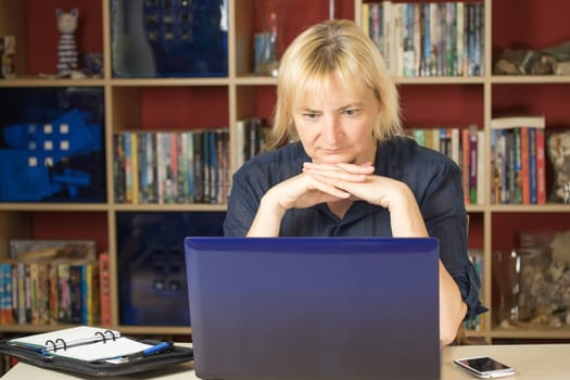 The middle aged blonde woman is reading intently at a notebook i home office. She is supporting her chin by both hands.. All potential trademarks are removed.