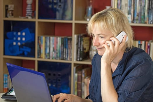 The middle aged blonde woman is smiling and calling a mobile phone. At the same time it is looking into the notebook. All potential trademarks are removed.