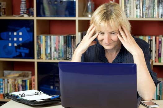 The middle aged woman is sitting at home and is working with a notebook. She is smiling and surprised by what she reads in the notebook. All potential trademarks are removed.
