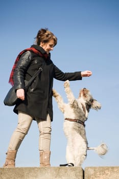 Full-body view of a mature woman with a mixed-breed dog standing on her hind legs in front of blue sky