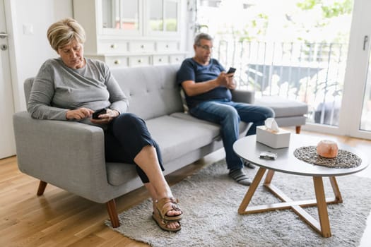 Senior couple sitting on couch and using their mobile phones at home