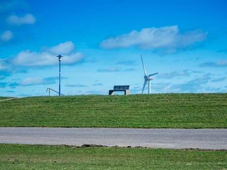 Landscape view of the German North Sea coast with a park bench on the dyke and a street lamp next to it, as well as a wind turbine in the background against a blue sky with a few clouds.