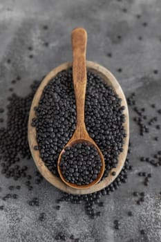 Bowl of dry black lentils beans with a spoon on grey table close up, copy space. Healthy eating, protein source for vegan and vegetarian diet. Traditional Asian and North African cuisines ingredient