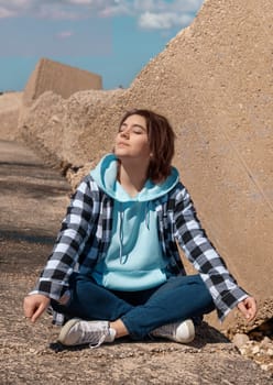 Happy Teenager girl in light blue hoodie and plaid shirt. Smiling Teen girl sitting outdoor cross-legged and enjoying the sunny day. Adolescence concept