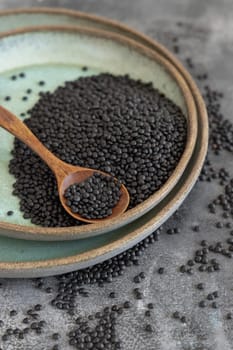 Plate of dry black lentils beans with a spoon on grey table close up. Healthy eating, protein source for vegan and vegetarian diet. Traditional Asian and North African cuisines ingredient