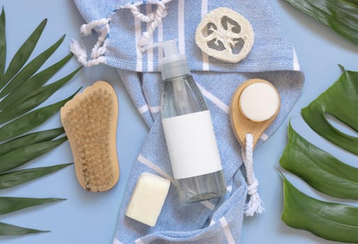 Transparent one pump dispenser, skin and hair care accessories on blue bath towel near tropical leaves top view, copy space. Packaging mockup. Natural homemade products