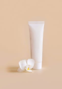White plastic cream tube near white orchid flower on light yellow, close up, mockup. Skincare beauty product, cream or lotion. Exotic natural cosmetics, pastel minimal composition..
