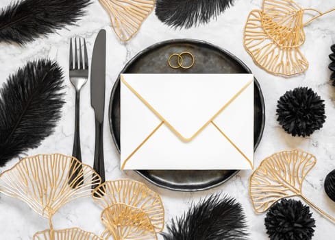 Black and golden wedding table setting with a blank envelope and rings top view, mockup. Elegant flat lay with horizontal greeting or Invitation paper envelope, wedding or holiday stationery template