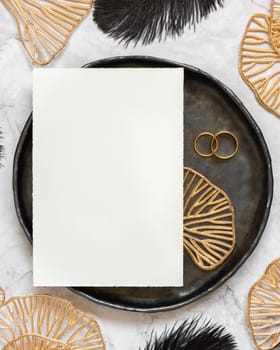 Black and golden wedding table setting with a blank card and rings top view, mockup. Elegant flat lay with vertical greeting or Invitation paper card, wedding or holiday stationery template