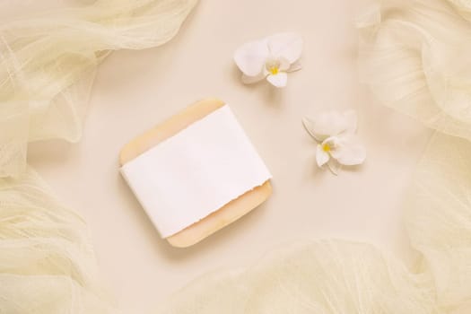 Hanmade soap bar near white orchid flowers and tulle on light yellow, top view, mockup. Skincare beauty product. Exotic natural cosmetics, pastel minimal composition..