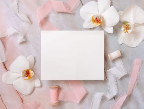 Wedding Card near white orchid flowers and silk ribbons on marble top view, mockup. Romantic scene with blank card flat lay, place for text. Valentines or Spring romantic concept