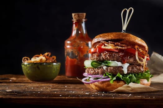Appetizing burger with cutlets and vegetable served on wooden table with ketchup and bowl of caramelized onion on black background