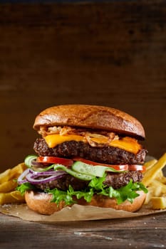 Delicious double burger with cheese on cutlets and vegetables served on parchment paper on wooden table with fries against brown background