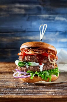 Tasty double burger with cutlets and assorted vegetables on lettuce served on parchment paper on table in room against wooden background