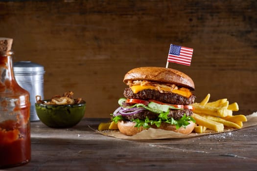 Appetizing burger with American flag skewer served on parchment paper on table with bottle of sauce and bowl of fried onions