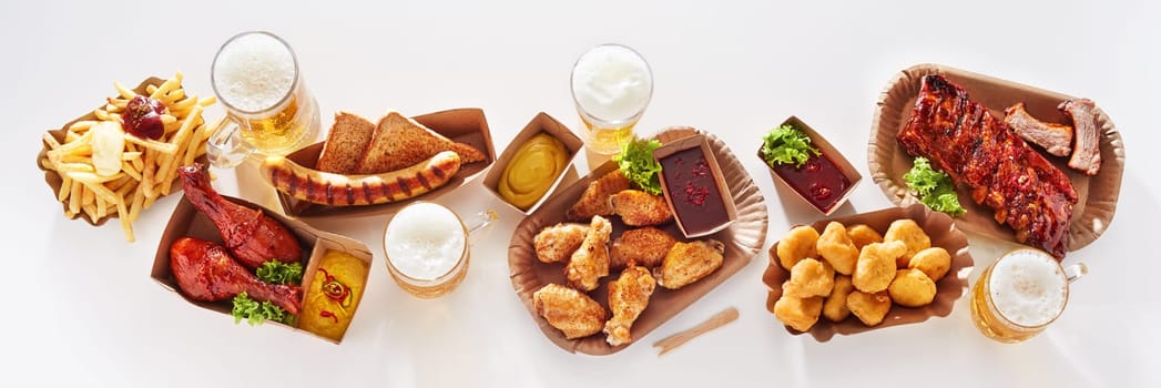 Top view of glass cups of lager beer served on white table with various meat and fast food in studio